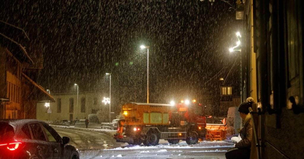 a snow plow truck driving along a snowy city street at night with lights, buildings, and traffic nearby.