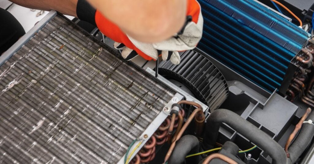 A gloved hand using a screwdriver inside an open air conditioning unit with coils, fan blades, wires, and tubing.