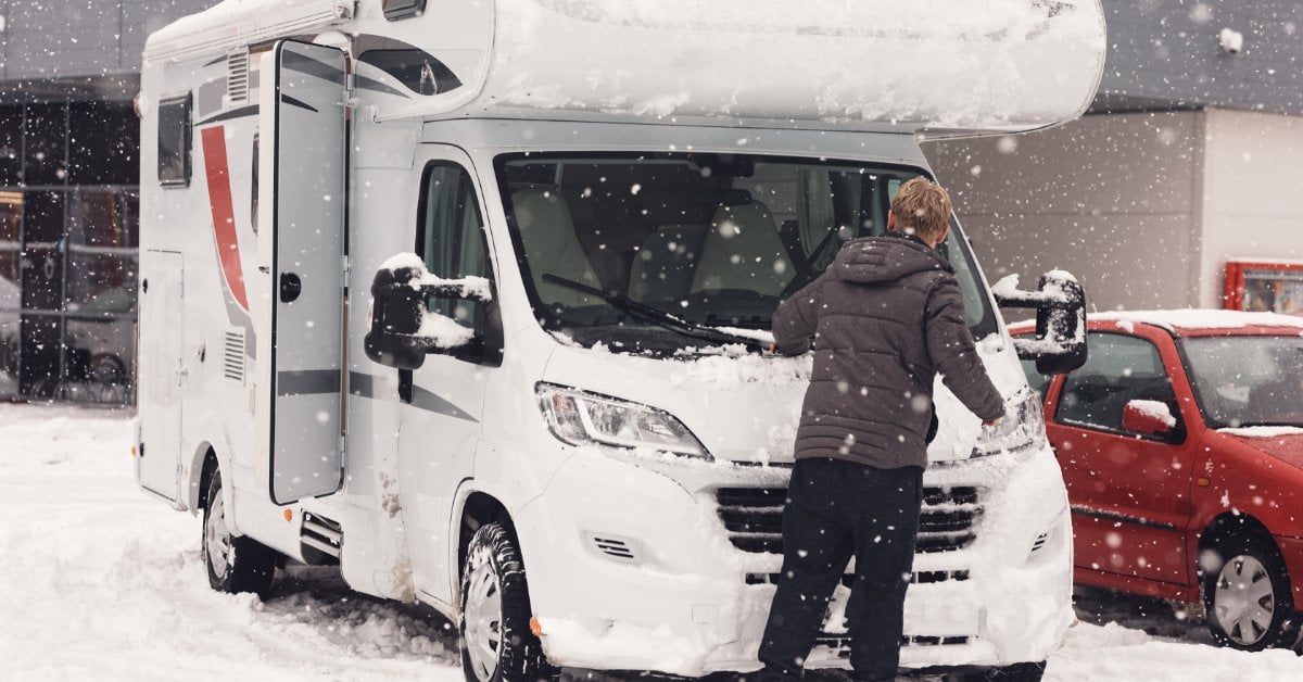 A person clearing snow from the front of a white camper van in a snowy parking lot beside a red car and building.