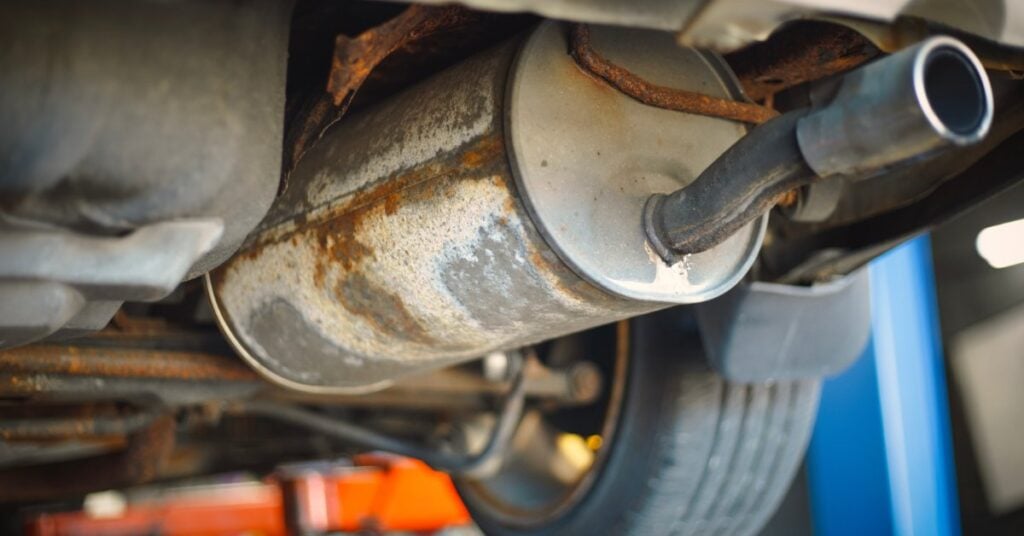A rusty exhaust pipe and muffler under a raised vehicle near a wheel, with blue lift equipment in the background.