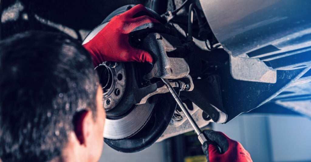 A person wearing red gloves using a hand tool on an exposed wheel hub and brake parts beneath a raised vehicle.